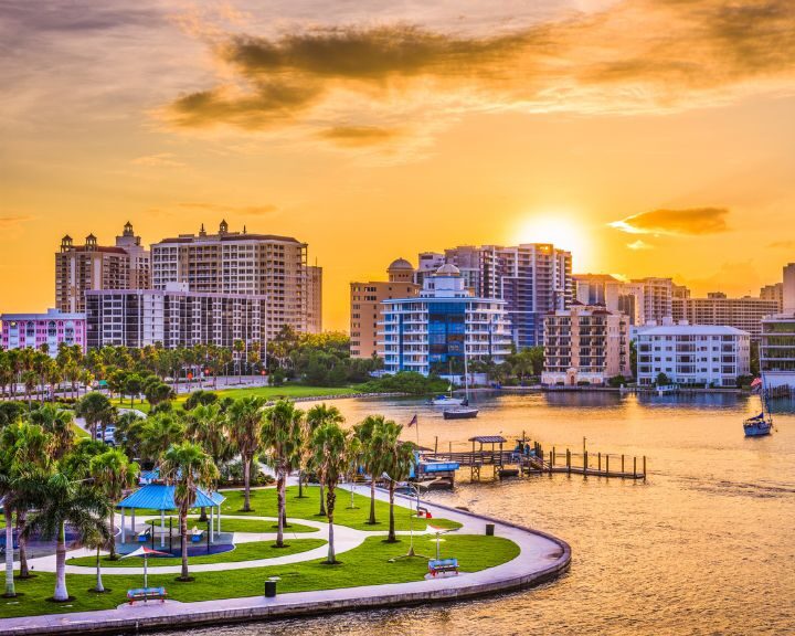The sun is setting over a city with palm trees and a waterway.